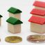 Drei Münten liegen auuf einer weißen Fläche, dahinter verschiedene Spielzeughäuser aus Holz mit grünen und roten Dächern