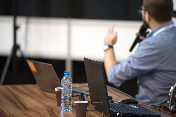 Ein Mann mit blauem Hemd sitzt an einem Konferenztisch mit Notebooks, hält ein Mikro in der Hand. Auf dem Tisch eine Wasserflasche